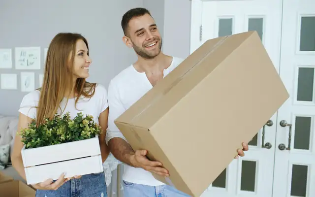A couple carrying a moving box and a potted plant