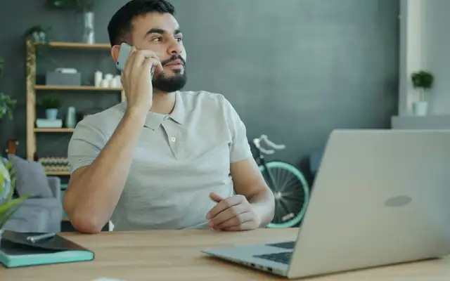 A man talking on his phone at a desk with an open laptop